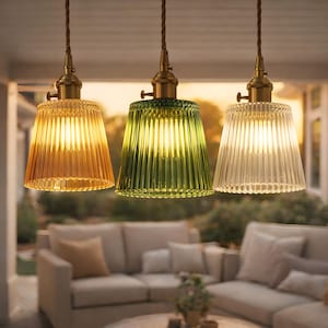 Könnte beinhalten: Drei Pendelleuchten mit gerippten Glasschirmen in Bernstein, Grün und Klar. Jede Leuchte hat eine Messingarmatur und ein braunes Kabel. Die Leuchten hängen über einer Terrasse mit einem Sofa.