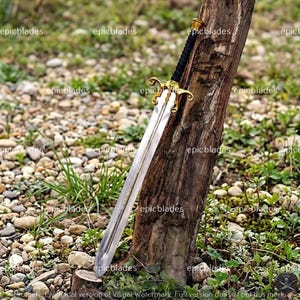 May include: A metal sword with a double-edged blade, a gold-colored ornate crossguard, and a black wrapped handle. The sword is positioned against a vertical tree trunk. The blade features a central fuller running down the length of the steel.