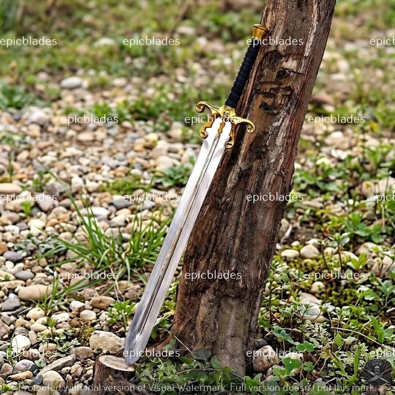 May include: A metal sword with a double-edged blade, a gold-colored ornate crossguard, and a black wrapped handle. The sword is positioned against a vertical tree trunk. The blade features a central fuller running down the length of the steel.