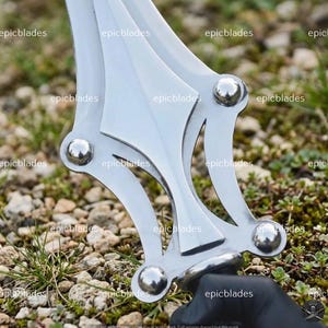 May include: A silver-colored metal sword guard featuring a symmetrical, open-work design with four prominent, polished spherical rivets at the corners. The metallic surface is reflective and smooth, showcasing a complex, geometric structure.