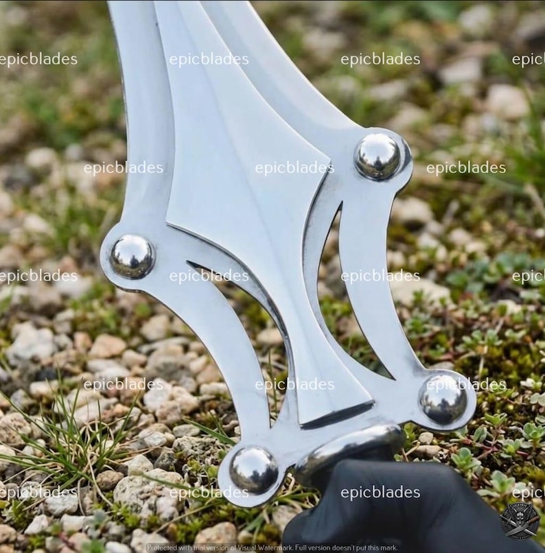 May include: A silver-colored metal sword guard featuring a symmetrical, open-work design with four prominent, polished spherical rivets at the corners. The metallic surface is reflective and smooth, showcasing a complex, geometric structure.