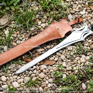 May include: A silver-colored metal sword with a black handle and a complex guard design, accompanied by a tan leather sheath featuring stitched edges and a metal ring attachment. The word epicblades is visible as a watermark across the image.