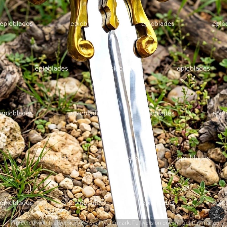 May include: A silver-colored metal sword blade with a central fuller groove, featuring a polished finish. The hilt area displays ornate, curved, gold-colored metal crossguard elements with scroll-like details. The text epicblades is visible.