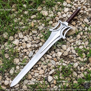 May include: A silver-colored metal sword featuring a double-edged blade, a complex open-work crossguard, and a dark brown leather-wrapped handle with a metallic pommel. The sword is positioned diagonally across a bed of small, light-colored stones.