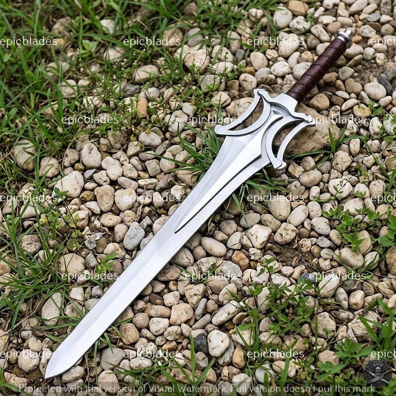 May include: A silver-colored metal sword featuring a double-edged blade, a complex open-work crossguard, and a dark brown leather-wrapped handle with a metallic pommel. The sword is positioned diagonally across a bed of small, light-colored stones.