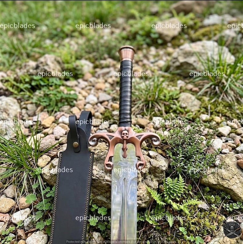 May include: A sword with a metallic blade, a black wrapped handle, and a copper-toned crossguard featuring a small skull detail at the center. The sword is accompanied by a black leather sheath with a metal snap closure and visible stitching.