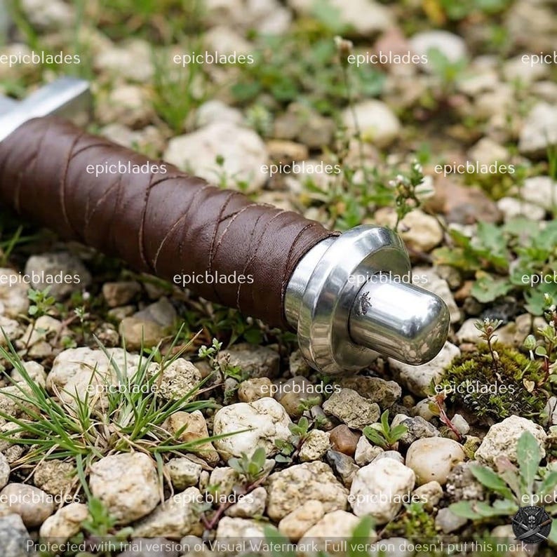May include: A sword handle wrapped in dark brown leather with a polished silver-colored metal pommel. The pommel features a small engraved emblem. The handle is positioned horizontally, showcasing the textured leather grip and metallic end cap.