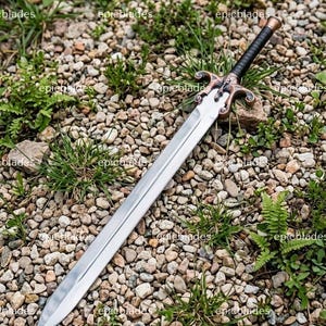 May include: A double-edged sword with a polished silver-colored steel blade, a black wrapped handle, and a decorative bronze-toned crossguard featuring a small skull emblem. The sword is positioned diagonally across a bed of small grey stones.