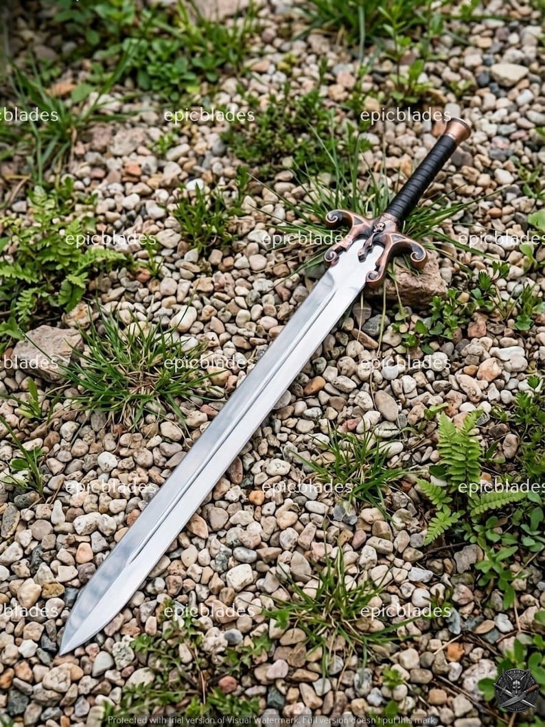 May include: A double-edged sword with a polished silver-colored steel blade, a black wrapped handle, and a decorative bronze-toned crossguard featuring a small skull emblem. The sword is positioned diagonally across a bed of small grey stones.