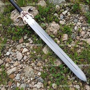 May include: A double-edged sword with a long, silver-toned steel blade featuring a central fuller. The hilt has a black wrapped grip, a copper-colored pommel, and a decorative crossguard with a central skull motif and curved quillons.
