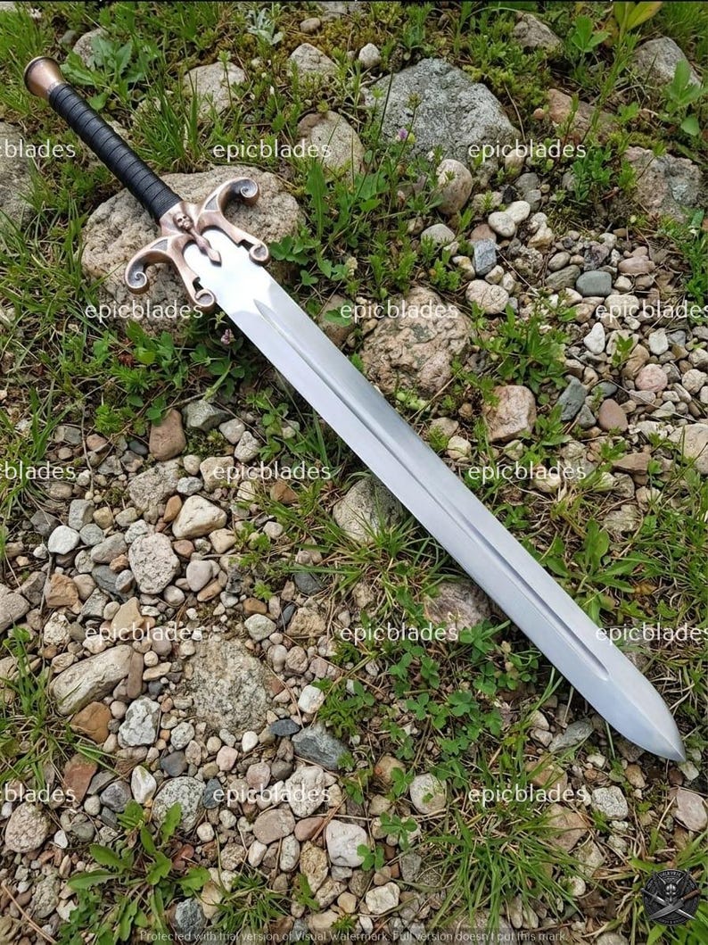 May include: A double-edged sword with a long, silver-toned steel blade featuring a central fuller. The hilt has a black wrapped grip, a copper-colored pommel, and a decorative crossguard with a central skull motif and curved quillons.