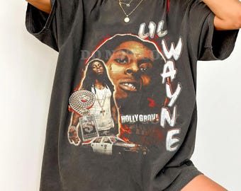 May include: Dark gray t-shirt featuring a graphic of Lil Wayne with the text "LIL WAYNE" in white. The design includes two images of the rapper and the words "HOLLY GROVE!" in red. The shirt has short sleeves and a relaxed fit.