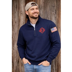 Könnte beinhalten: Marineblaues Sweatshirt mit Viertelreißverschluss, rotem Monogramm und einem amerikanischen Flaggenaufnäher am Ärmel. Das Sweatshirt hat einen Stehkragen und einen Reißverschluss. Die Person trägt eine Baseballkappe und Jeans.