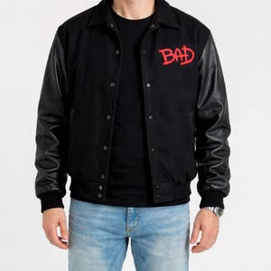May include: Black varsity jacket with black leather sleeves and a red "BAD" logo on the chest. The jacket has a button-up front, a collar, and ribbed cuffs and hem. The person is wearing blue jeans.