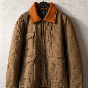 May include: A brown quilted jacket with an orange collar hangs on a black hook. The jacket has a zipper closure and four front pockets. The jacket is made of a durable material and is suitable for cooler weather.