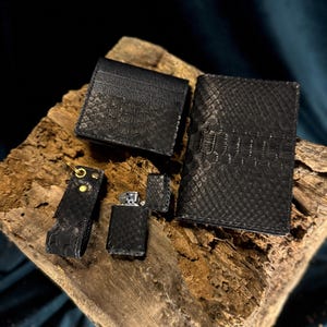 Peut inclure: Ensemble d'accessoires en cuir noir, comprenant un portefeuille, un porte-cartes, un briquet, un porte-clés et un porte-passeport, tous avec un motif texturé en peau de serpent. Les articles sont disposés sur un morceau de bois.