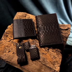 Könnte beinhalten: Eine Kollektion schwarzer Lederaccessoires, darunter ein Portemonnaie, ein Kartenetui, ein Feuerzeug und ein Schlüsselanhänger, alle mit einem strukturierten Schlangenmuster. Die Artikel werden auf einem Holzstück präsentiert, das die Handwerkskunst zeigt.