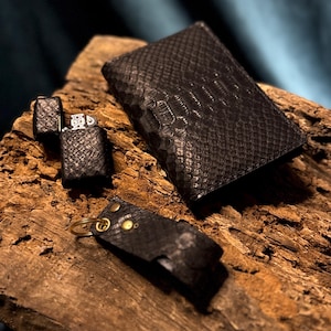 May include: A set of black leather accessories, including a lighter, a wallet, and a keyring holder, all featuring a textured, snake-skin pattern. The items are displayed on a piece of weathered wood, creating a rustic and stylish presentation.