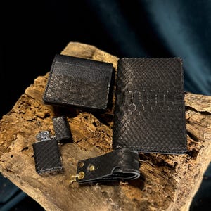 Könnte beinhalten: Eine Sammlung schwarzer Lederaccessoires, darunter ein Kartenhalter, eine Geldbörse, ein Feuerzeug und ein Schlüsselhalter, alle mit einem strukturierten Muster. Die Artikel werden auf einem Holzstück präsentiert, das die Handwerkskunst und das Design zeigt.