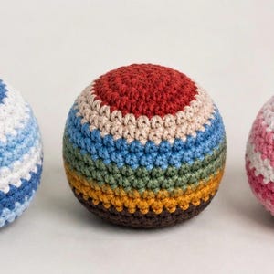 May include: Three colorful, crocheted balls. One is blue and white, another is a mix of red, tan, blue, green, and brown, and the third is pink and white. The balls are round and appear to be handmade.