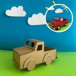 Puede incluir: Una camioneta de cartón marrón con ruedas negras. La camioneta está colocada sobre un fondo azul con nubes de papel blanco y una camioneta roja en un círculo. La camioneta es un proyecto de manualidades.