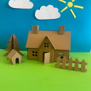 May include: A handmade cardboard house, tree, dog house, fence, sun, and clouds against a blue and green background. The house features a pitched roof, chimney, and windows. The sun is yellow, and the clouds are white.