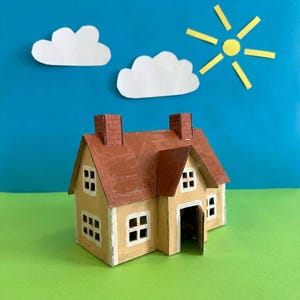 May include: A handmade miniature house made of cardboard, painted in beige with a brown roof and chimneys. The house has small square windows and an open door. The background features paper cutouts of clouds and a sun against a blue sky.