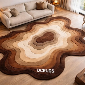 May include: A large, abstract area rug with a layered, organic design. The rug features a gradient of brown, tan, and cream colors, creating a warm and inviting aesthetic. The rug is placed on a hardwood floor, and the text "DCRUGS" is visible.