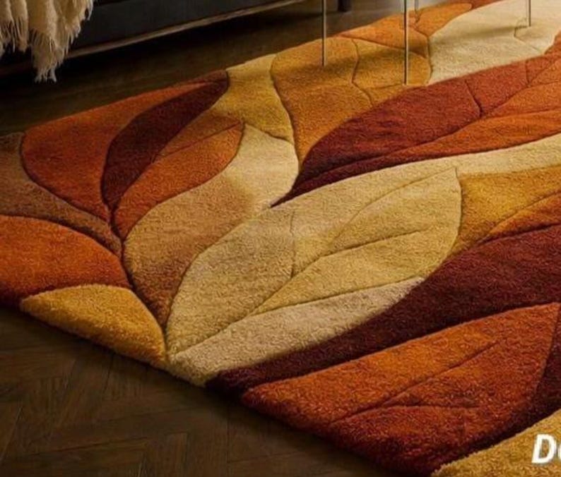 The rug is placed in a sophisticated living room setting with dark herringbone wood flooring that contrasts beautifully with the warm tones of the carpet. A modern charcoal gray sofa sits to the left, styled with neutral and olive green throw pillows and a soft cream knit blanket draped casually over the arm. In front of the sofa is a sleek glass coffee table with minimalist metal legs, allowing the bold rug pattern to remain fully visible underneath. The transparent table surface keeps the room