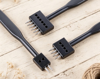 May include: Three black leathercraft tools with varying numbers of prongs are arranged on a light wood surface. The tools have a handle and a rectangular head with sharp, pointed prongs for punching holes in leather. A ball of twine and dried flowers are also visible.