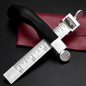 May include: A leather edge creaser tool with a black handle and silver-colored metal components. The tool features a ruler with inch markings and a screw for adjustments. The background includes a red leather surface.