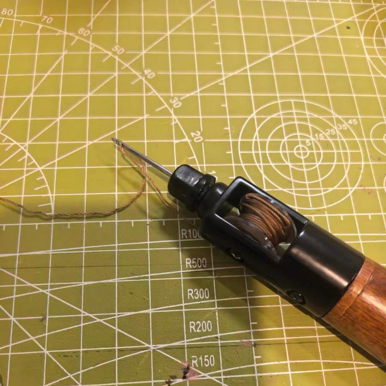 May include: A leather stitching awl with a wooden handle and a black metal body. The awl has a needle extending from the tip and a spool of thread inside. The tool is on a green cutting mat.