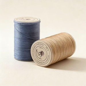 Puede incluir: Dos carretes de hilo de coser. Uno es azul claro, de pie. El otro es de color beige, tumbado de lado. Los carretes tienen etiquetas blancas con texto y un dibujo de máquina de coser. Ideal para costura.