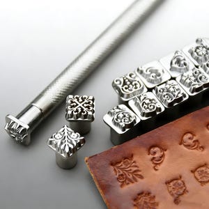 Pode incluir: Um conjunto de ferramentas de estampagem de couro em metal prateado com vários designs intrincados de flores e volutas. A coleção inclui um cabo longo e várias cabeças quadradas, com um pedaço de couro castanho mostrando impressões.