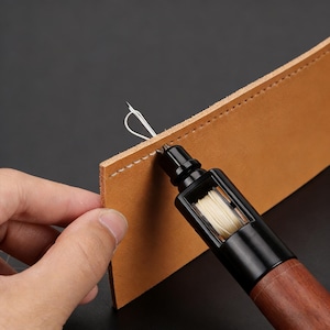 Puede incluir: Una herramienta de costura de cuero con mango de madera y cuerpo negro se utiliza. La herramienta está cosiendo un trozo de cuero marrón con hilo blanco. La aguja se inserta a través del cuero, demostrando el proceso de costura.