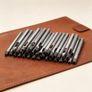 May include: A selection of silver metal hole punch tools with different shaped cutting ends. The tools are placed on a brown leather surface, indicating a craft or leatherworking focus. These tools are designed for creating precise shapes in materials.