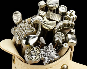 May include: A collection of metal stamps with various decorative designs, including flowers, geometric shapes, and textured patterns. The stamps are stored in a beige leather pouch against a black background.