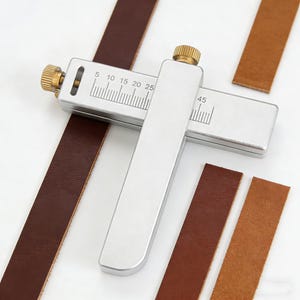 May include: A silver leather edge creaser tool with brass knobs, measuring in millimeters, is shown with brown leather strips. The tool is designed for precise leatherworking, with clear markings for accurate measurements.