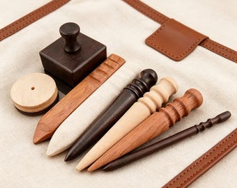 May include: A collection of leather crafting tools including a dark brown wooden stamp, a light wood circular disc, a flat wooden burnisher, and four tapered wooden burnishing sticks in varying shades of brown and light wood tones.