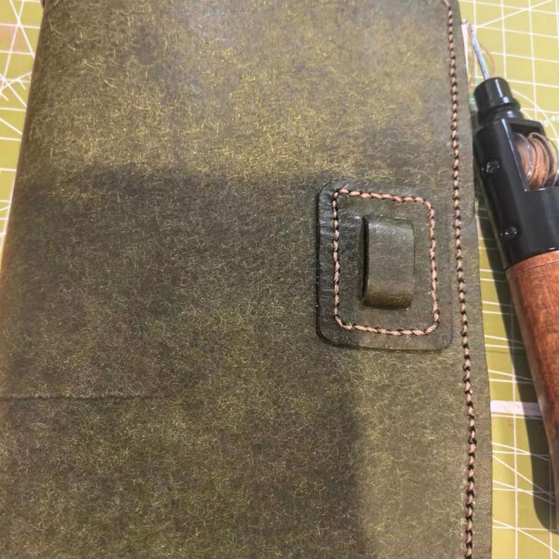May include: A handcrafted, olive green leather journal with a rectangular clasp and stitched edges. A leather crafting tool with a wooden handle and metal tip is positioned on the right side of the journal.