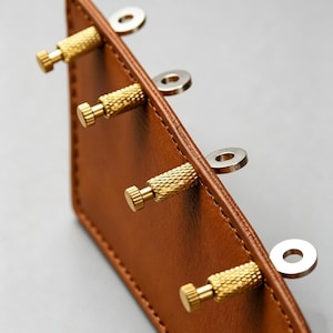 May include: Close-up of a brown leather item with four brass screws and silver washers. The leather has a stitched edge and a smooth surface. The brass screws have a textured grip.