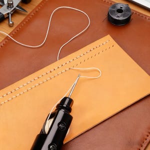 May include: A close-up of leather crafting tools. A black stitching awl with a needle is inserted into a piece of tan leather. White thread is visible, along with a spool of thread and a leather punch tool. The background is brown leather.