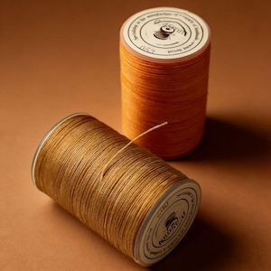 May include: Two spools of thread in shades of orange and gold. The spools have white labels with black text and a small illustration. One spool is lying on its side, with a strand of thread extending from it. The background is a solid brown.