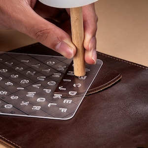 Puede incluir: Una plantilla transparente de letras y números de acrílico se coloca sobre una superficie de cuero marrón. Se utiliza un mazo de madera para estampar las letras y los números en el cuero. La plantilla incluye el alfabeto y los números del 1 al 12.