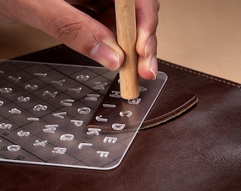 May include: A clear acrylic letter and number stamping template is positioned over a brown leather surface. A wooden mallet is used to stamp the letters and numbers onto the leather. The template includes the alphabet and numbers 1-12.