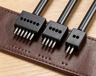 May include: Three black leather hole punch tools of varying sizes are positioned over a brown leather strap. The tools have multiple metal prongs for creating holes. The leather strap has a series of pre-punched holes.