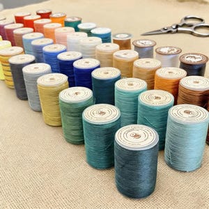 Pode incluir: Uma coleção de bobinas de linha coloridas dispostas sobre uma superfície bege. As bobinas são de várias cores, incluindo vermelho, azul, verde, amarelo e laranja. Uma tesoura prateada está no fundo.