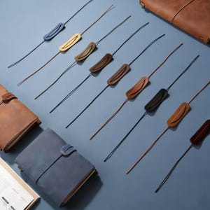 Puede incluir: Una variedad de marcadores de cuero para diarios en varios colores, incluyendo azul, marrón y negro, con diarios de cuero a juego. Los marcadores tienen una pieza rectangular de cuero unida a un cordón fino. Los diarios se cierran con una correa de cuero.