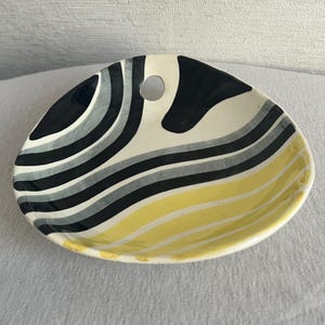Könnte beinhalten: Eine Keramikschale mit einer einzigartigen, asymmetrischen Form. Die Schale hat ein Design aus schwarzen, grauen und gelben gebogenen Streifen. Sie hat ein kleines Loch in der Nähe der Oberseite und dient wahrscheinlich zum Servieren oder Ausstellen.