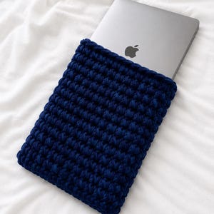 Op de afbeelding: Een donkerblauwe gehaakte laptophoes gemaakt van dik garen, met een dicht en gestructureerd steekpatroon. De rechthoekige hoes is aan de bovenkant open en bevat gedeeltelijk een zilverkleurige laptop met een zwart Apple-logo op het deksel.